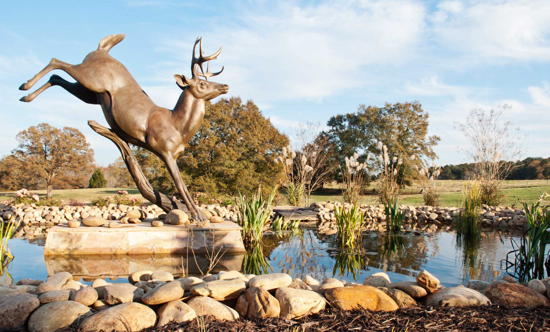 A deer sculpture standing on a manicured lawn next to a tranquil, man-made lagoon in a beautifully landscaped yard.